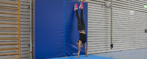 Jugendliche führt einen Handstand aus in einer Sporthallte an einer Prallschutzmatte an Sprossenwand